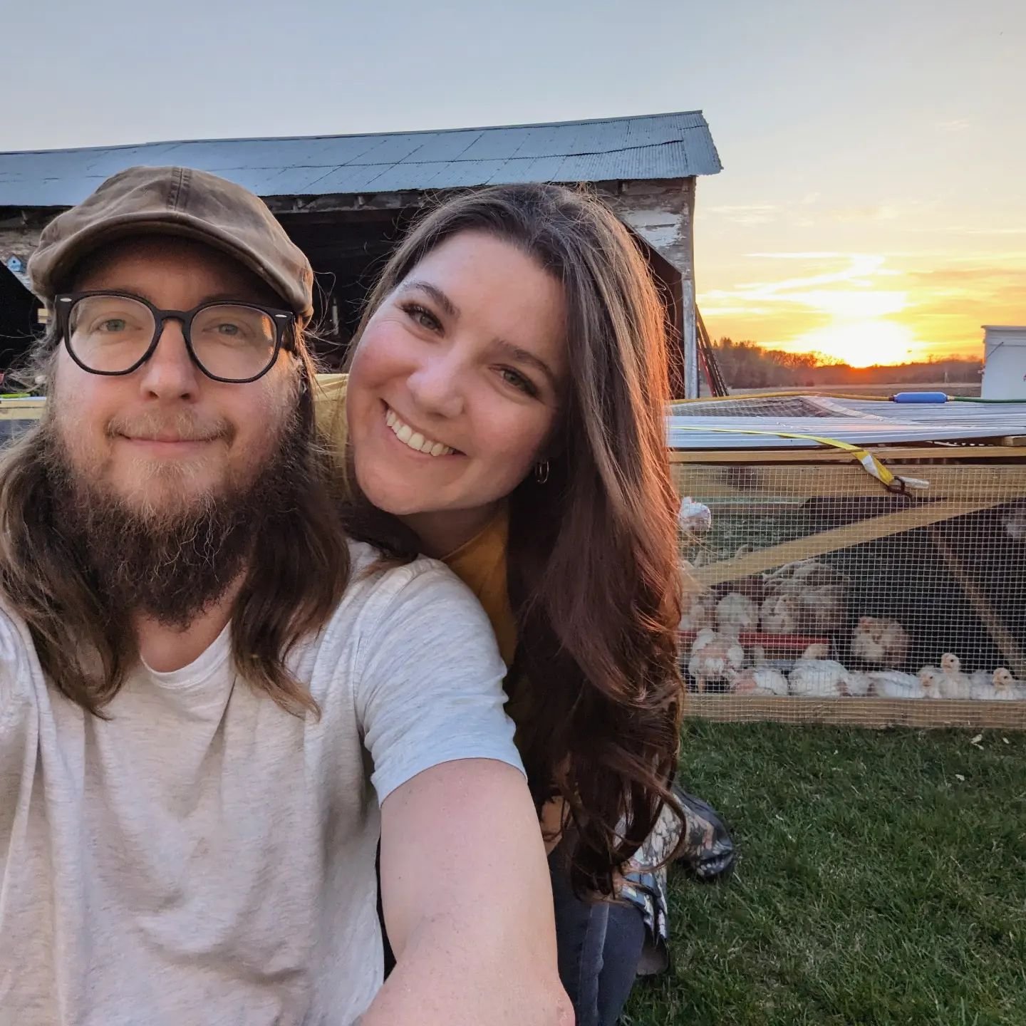 Couple posing for a selfie on a farm at sunset, with a chicken coop in the background, showcasing rural life and agriculture.