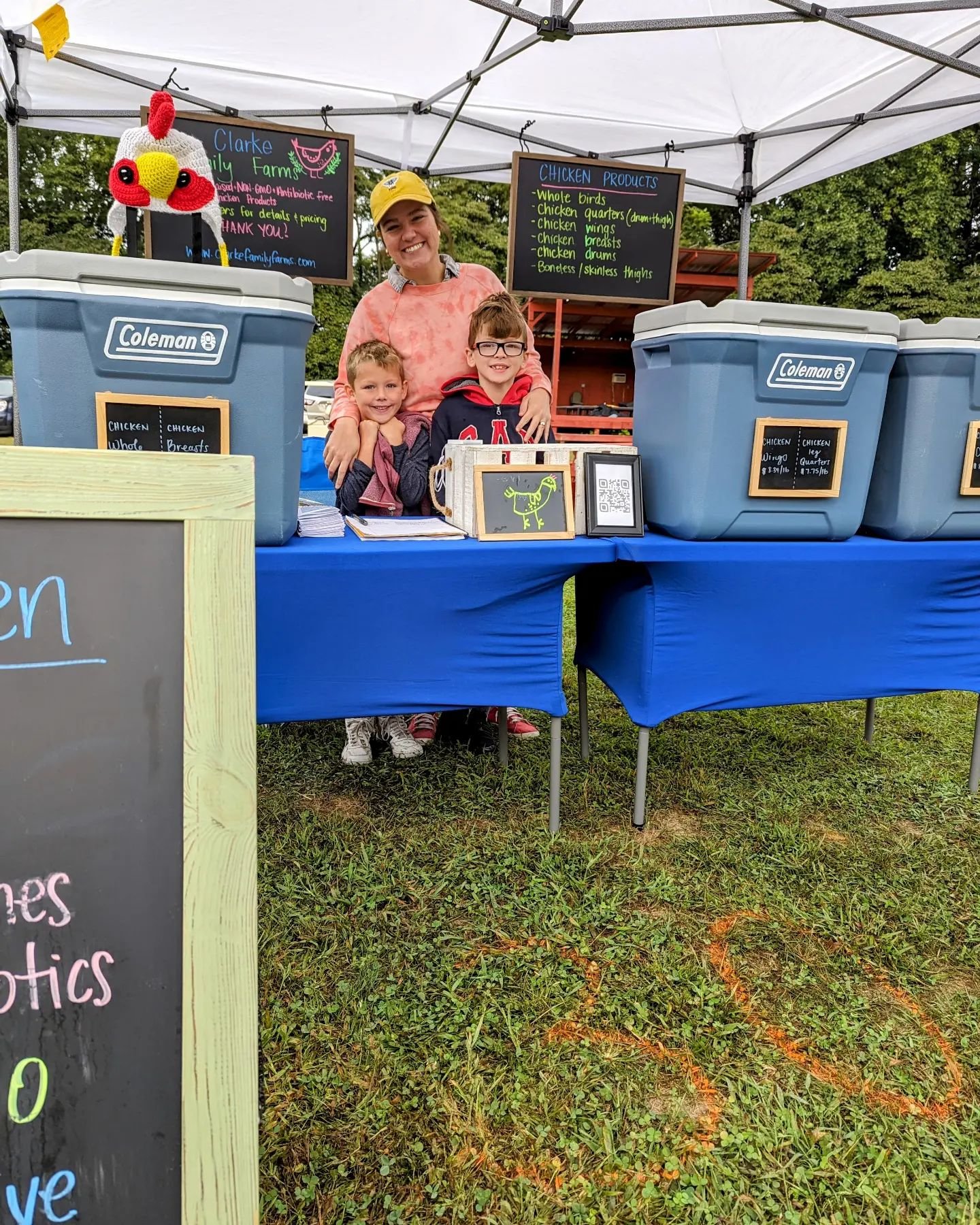 Family-run farm stand at a local market featuring chicken products, with a smiling woman and two children promoting their offerings. The display includes coolers labeled with various chicken products, while a chalkboard lists available items.