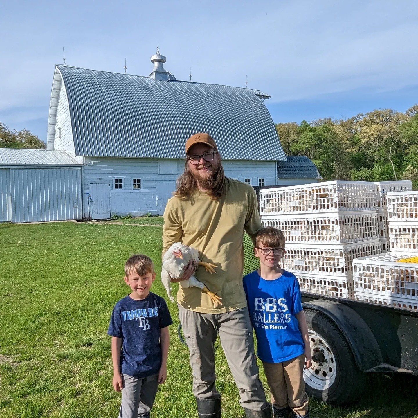 A farmer holding a chicken stands with two young boys in front of a barn and a trailer filled with crates. The scene captures a sunny day on a farm, showcasing rural life and family involvement in agriculture.