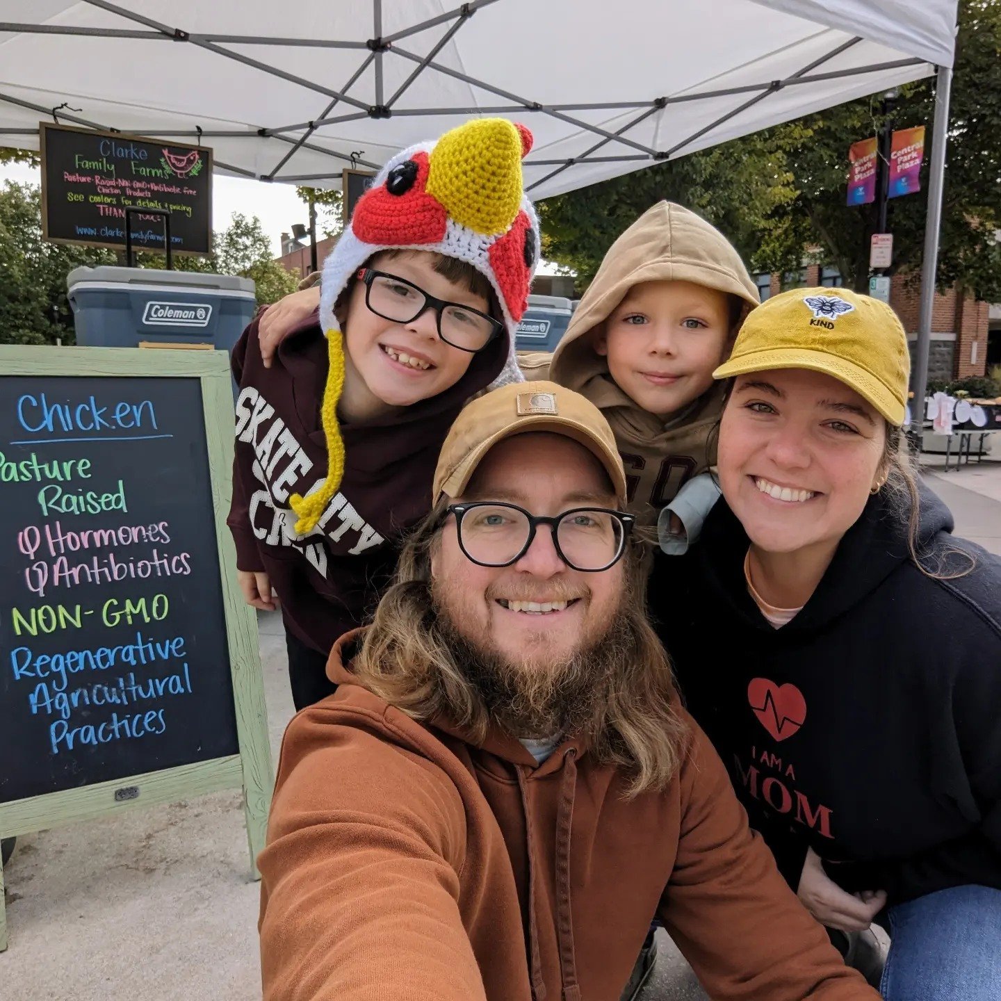 Family portrait at a farmer's market featuring a man with glasses and a beard, a woman smiling, and two children, one wearing a chicken hat. Behind them is a sign promoting pasture-raised, non-GMO chicken with no hormones or antibiotics, highlighting regenerative agricultural practices.