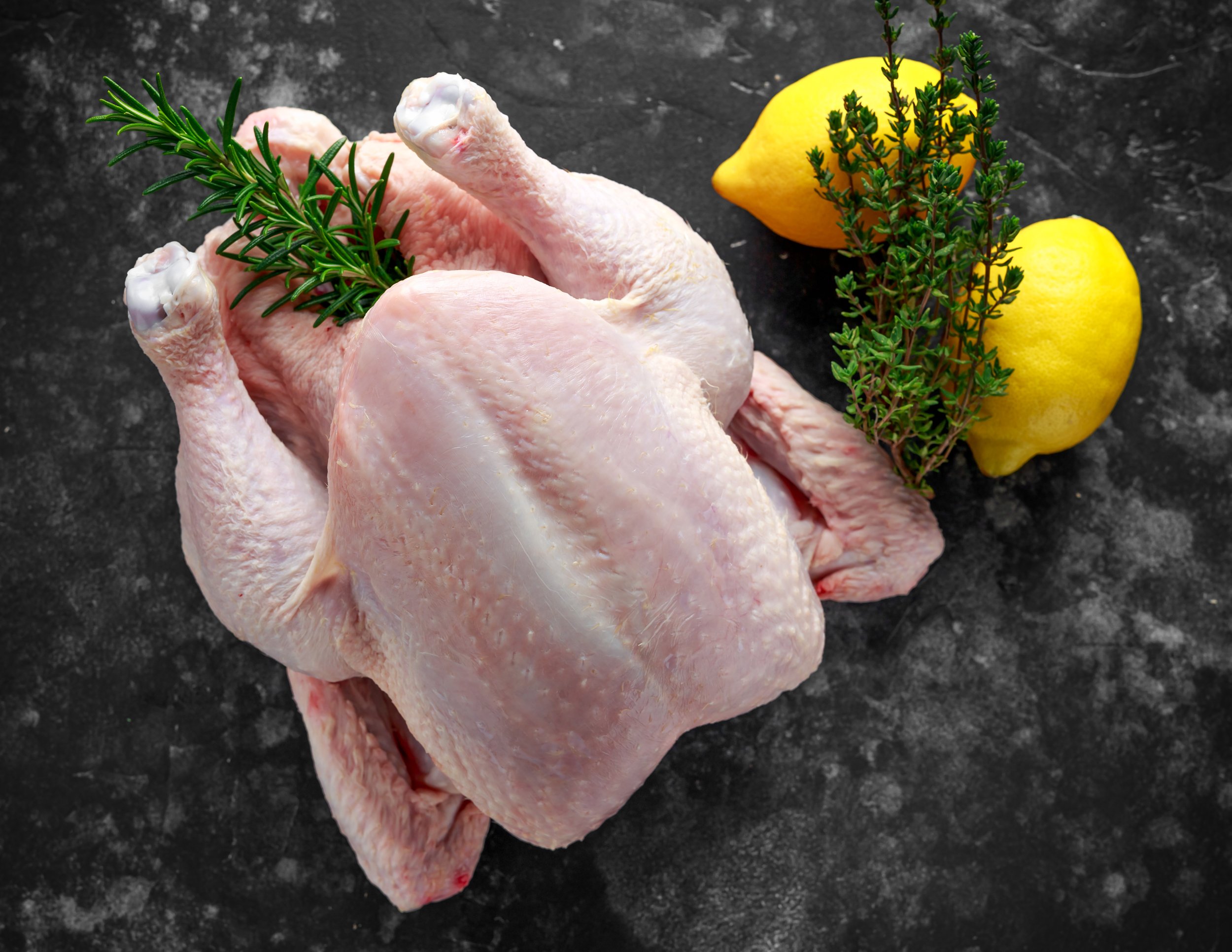 Raw whole chicken with rosemary and thyme, accompanied by two lemons, resting on a dark textured surface. Ideal for preparing flavorful dishes.